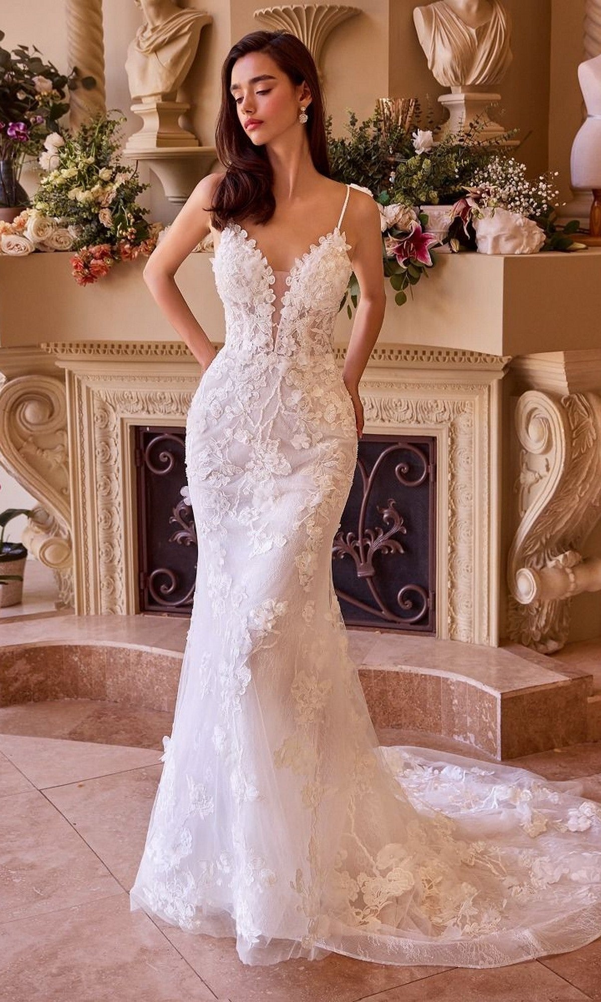A woman in the Andrea and Leo WL053 long wedding dress with floral lace applique and an adjustable lace-up back stands indoors by a fireplace adorned with flowers and classical sculptures.